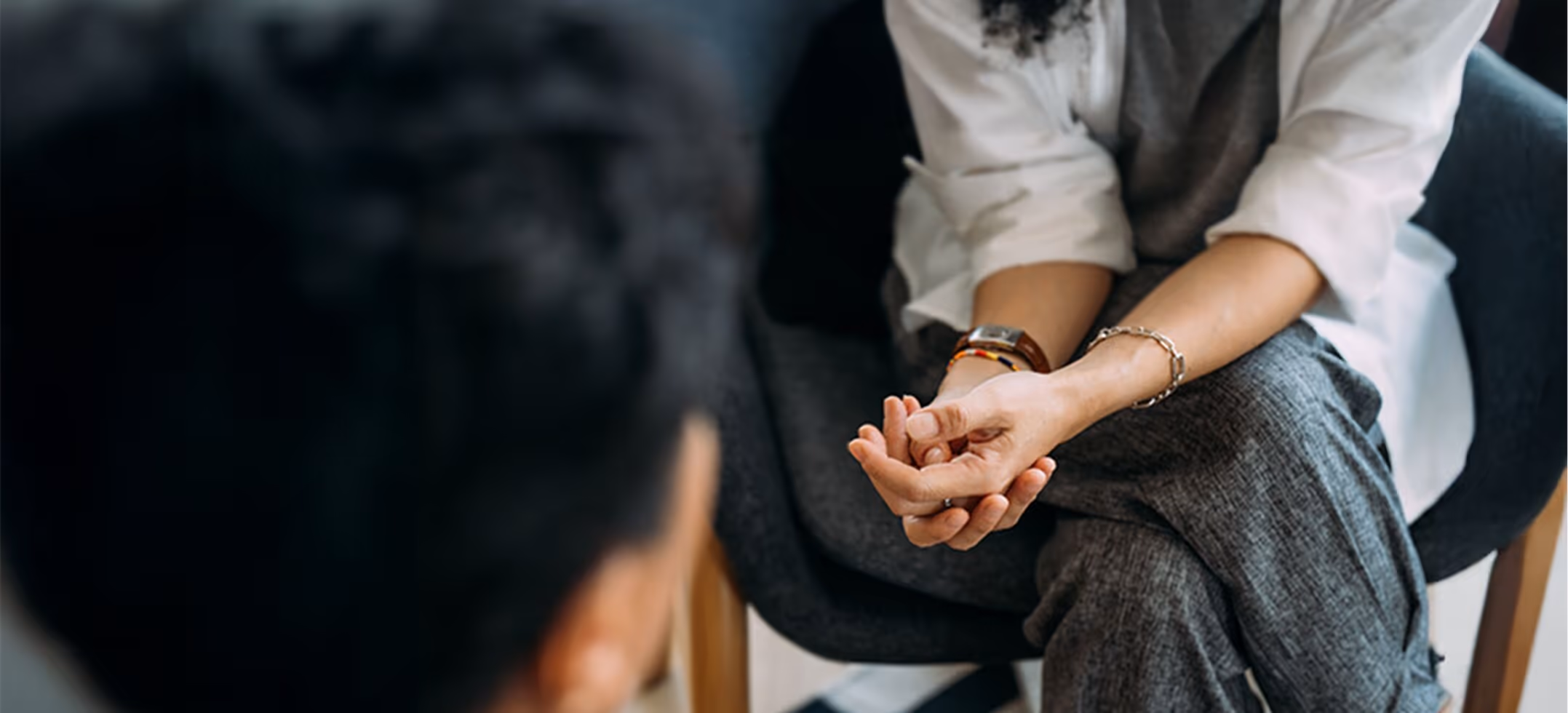 A therapist sitting with hands folded, seen past the shoulder of the client facing her