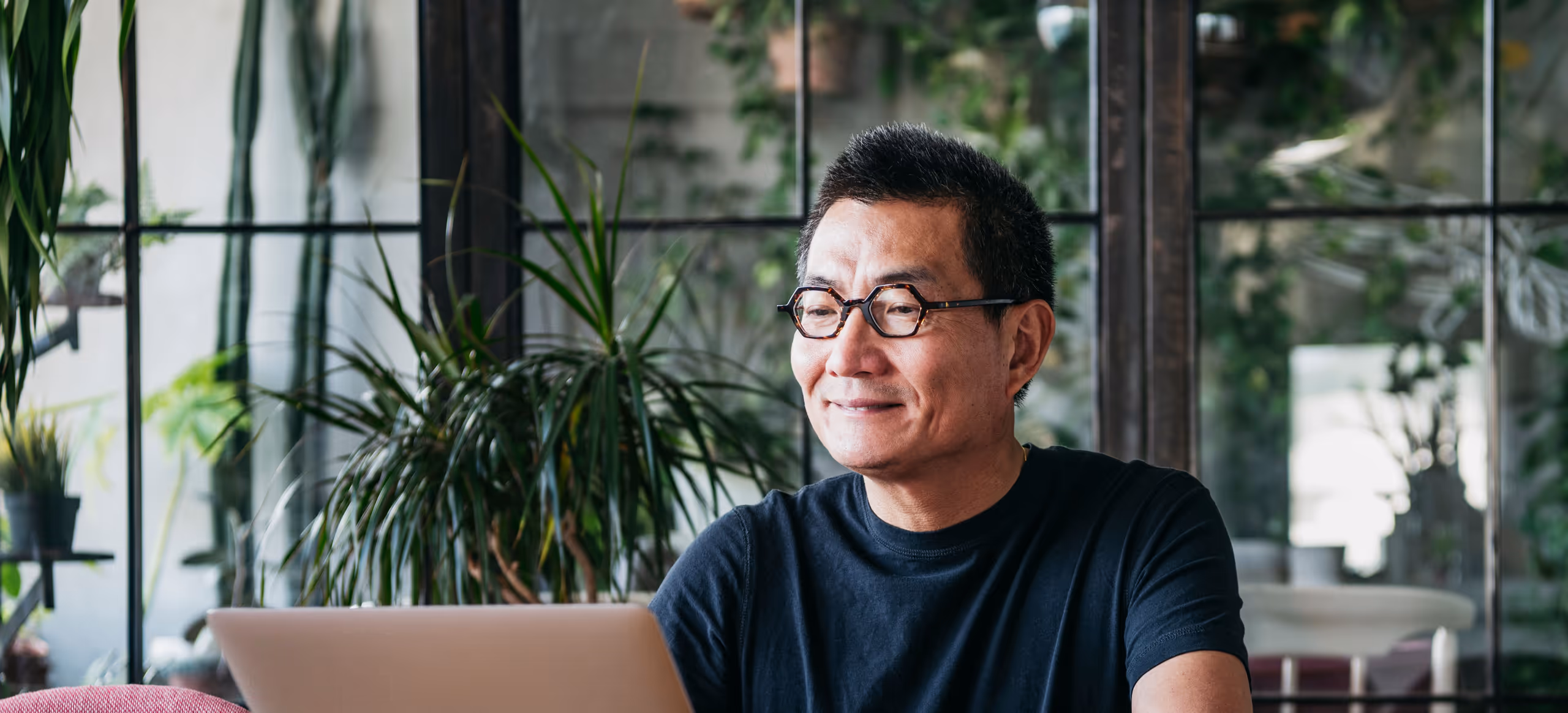 A man in glasses sitting at a laptop in a plant-filled room, looking out of a window