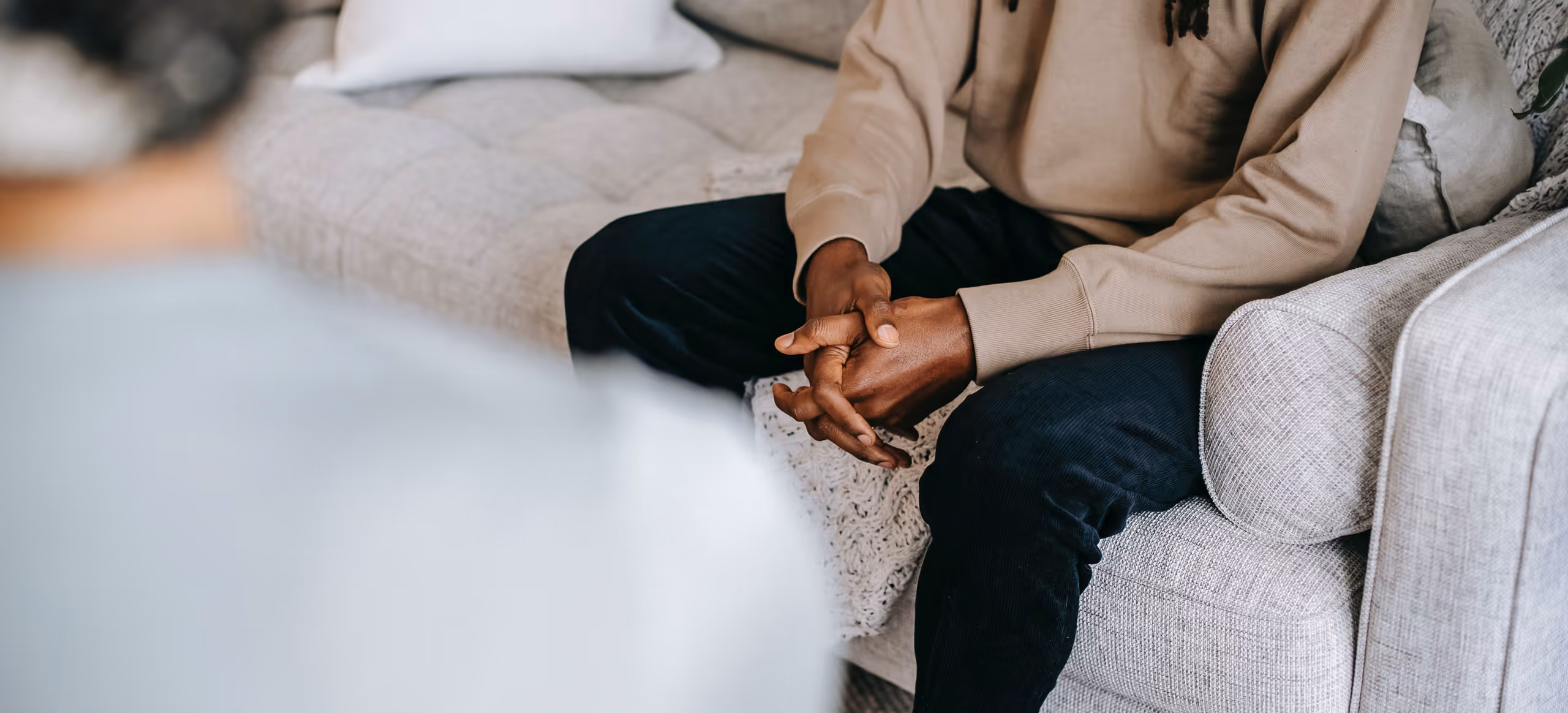 A man sitting forward on a couch with his hands clasped, a therapist out of focus in the foreground
