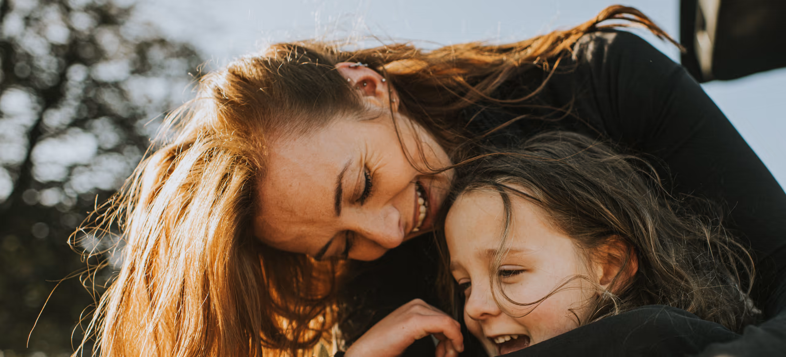 A mother and daughter laughing as they hug outdoors in the sunlight