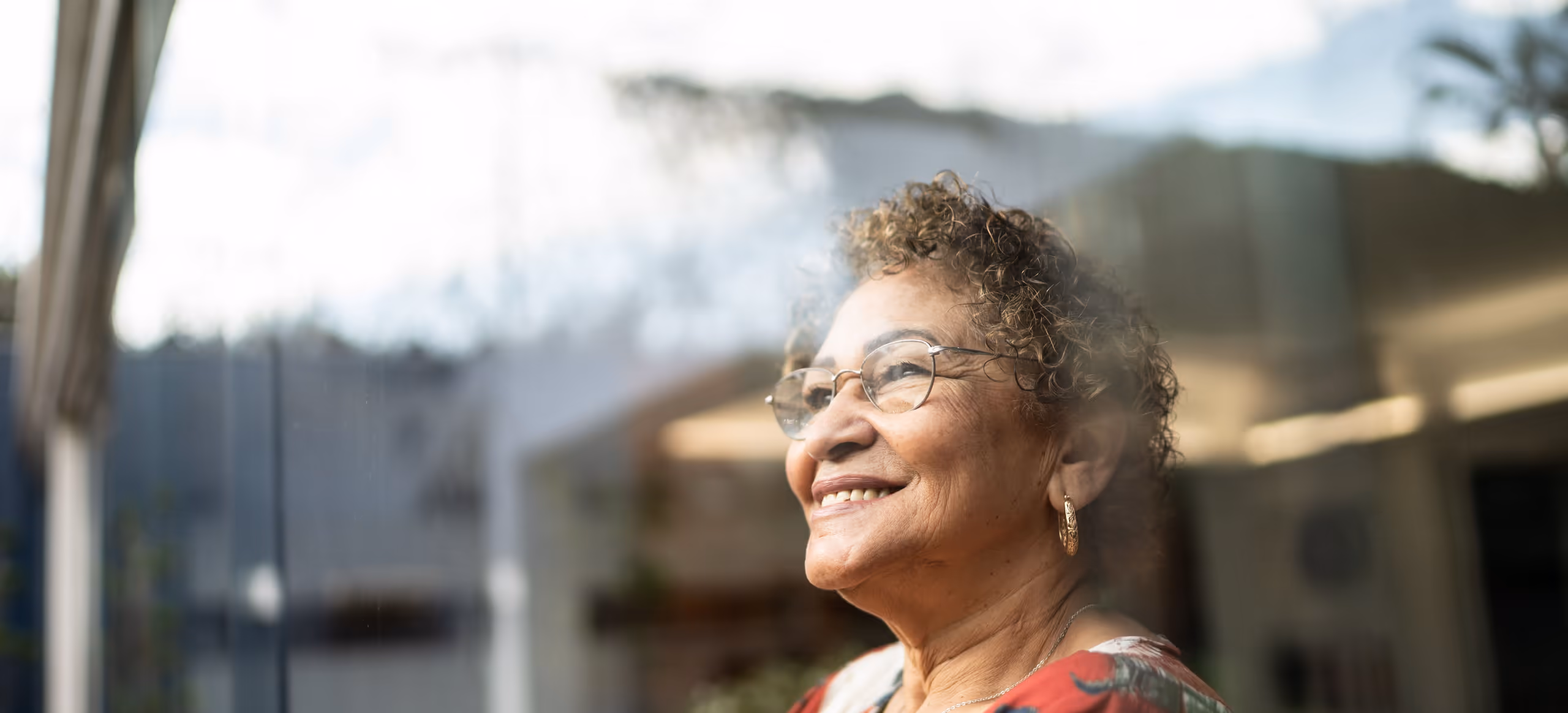 An older woman with grey curls and glasses smiling in the light of a window