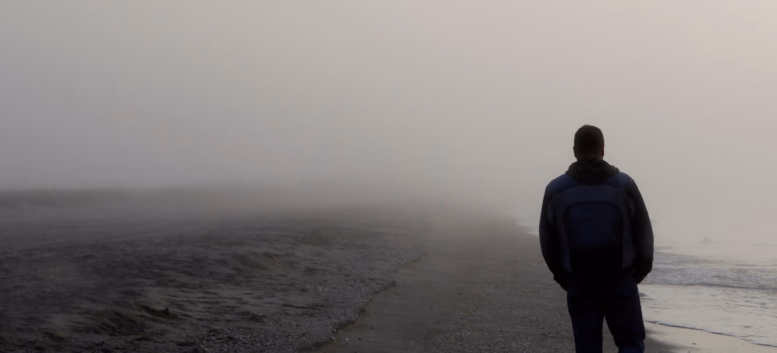 A lone figure walking along the edge of the water on a foggy beach
