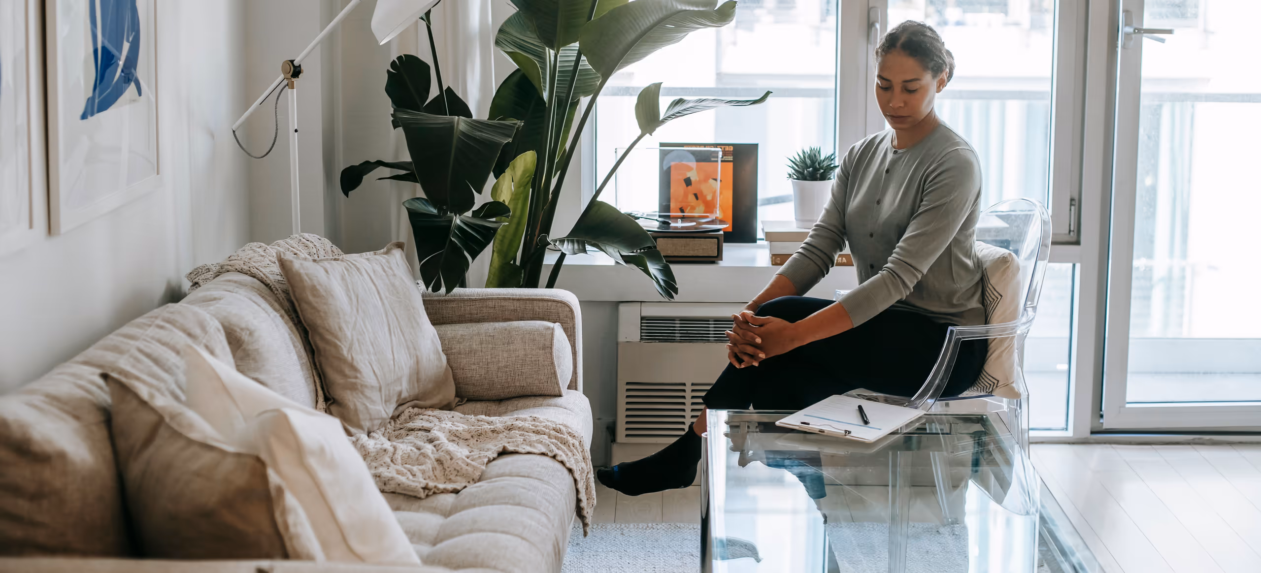 A therapist sitting forward in a chair in a sunlit room, a couch and notepad beside her