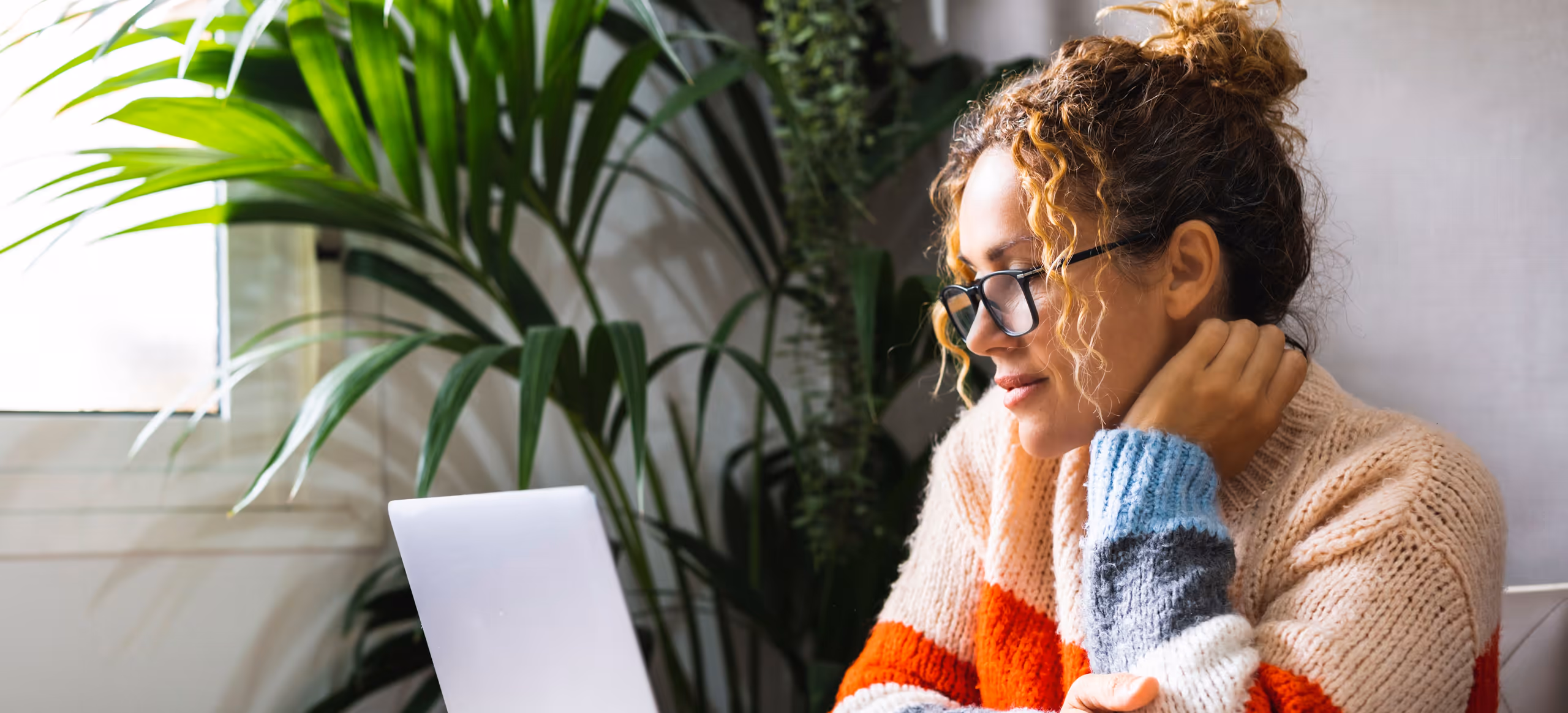 A woman in a knitted jumper at a laptop beside a houseplant, resting her chin on her hand