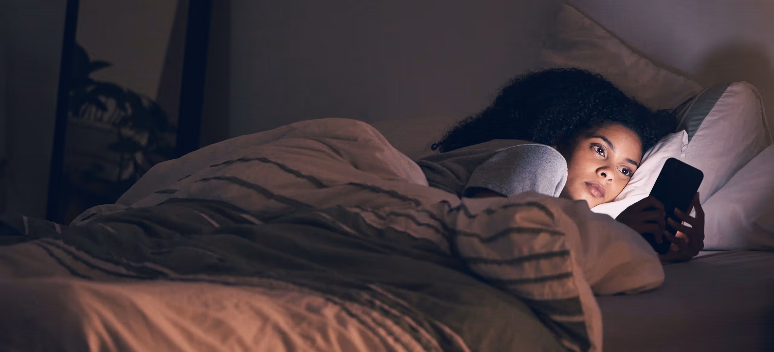 A woman lying awake in bed at night, her face lit by the phone in her hand