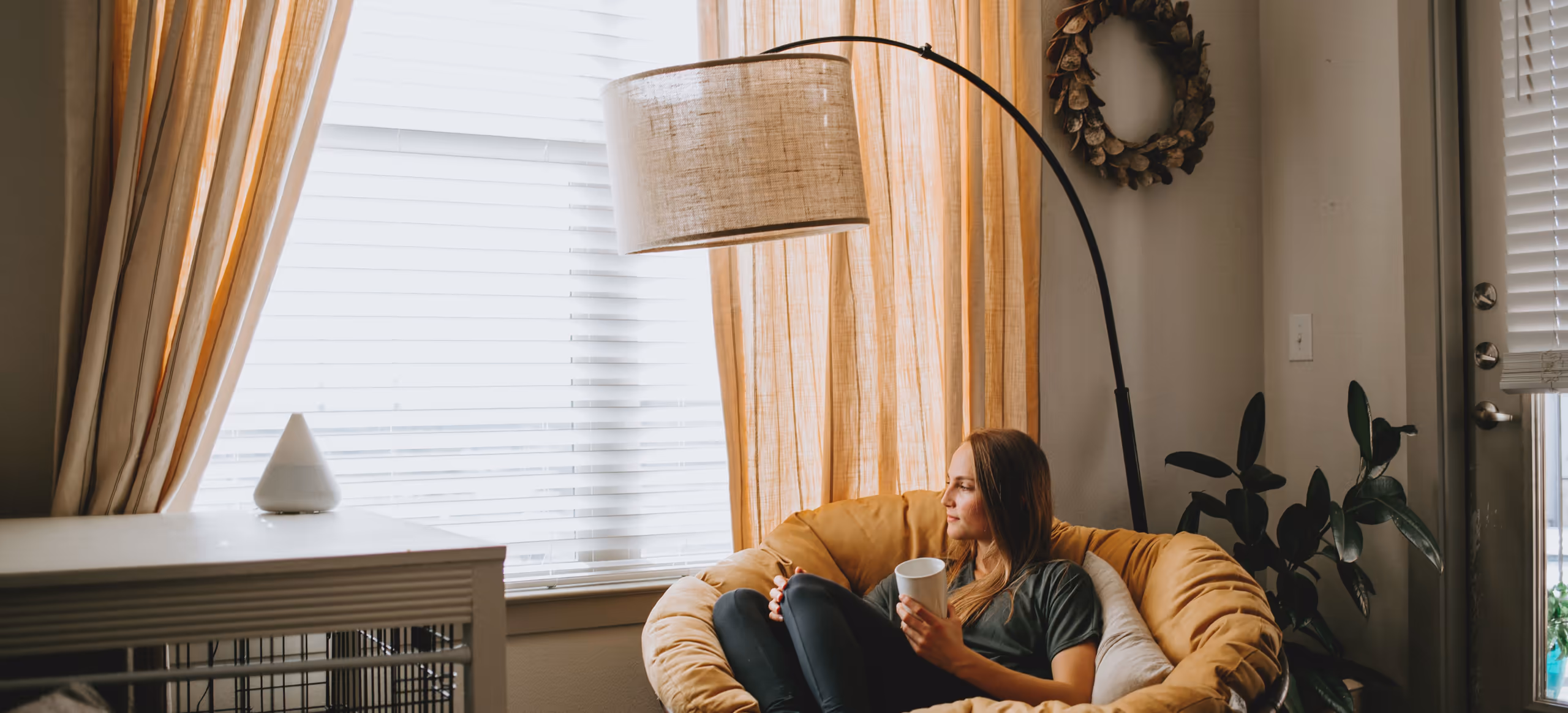 A woman curled into a round chair by a window, holding a mug