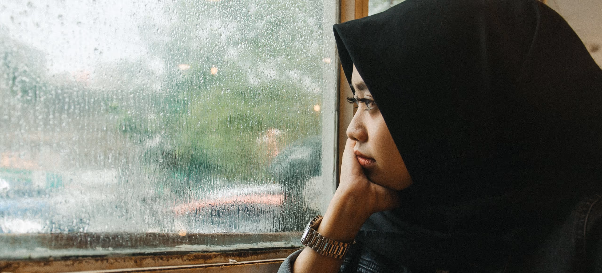 A woman in a hijab sitting by a rain-streaked cafe window with a coffee, looking outside