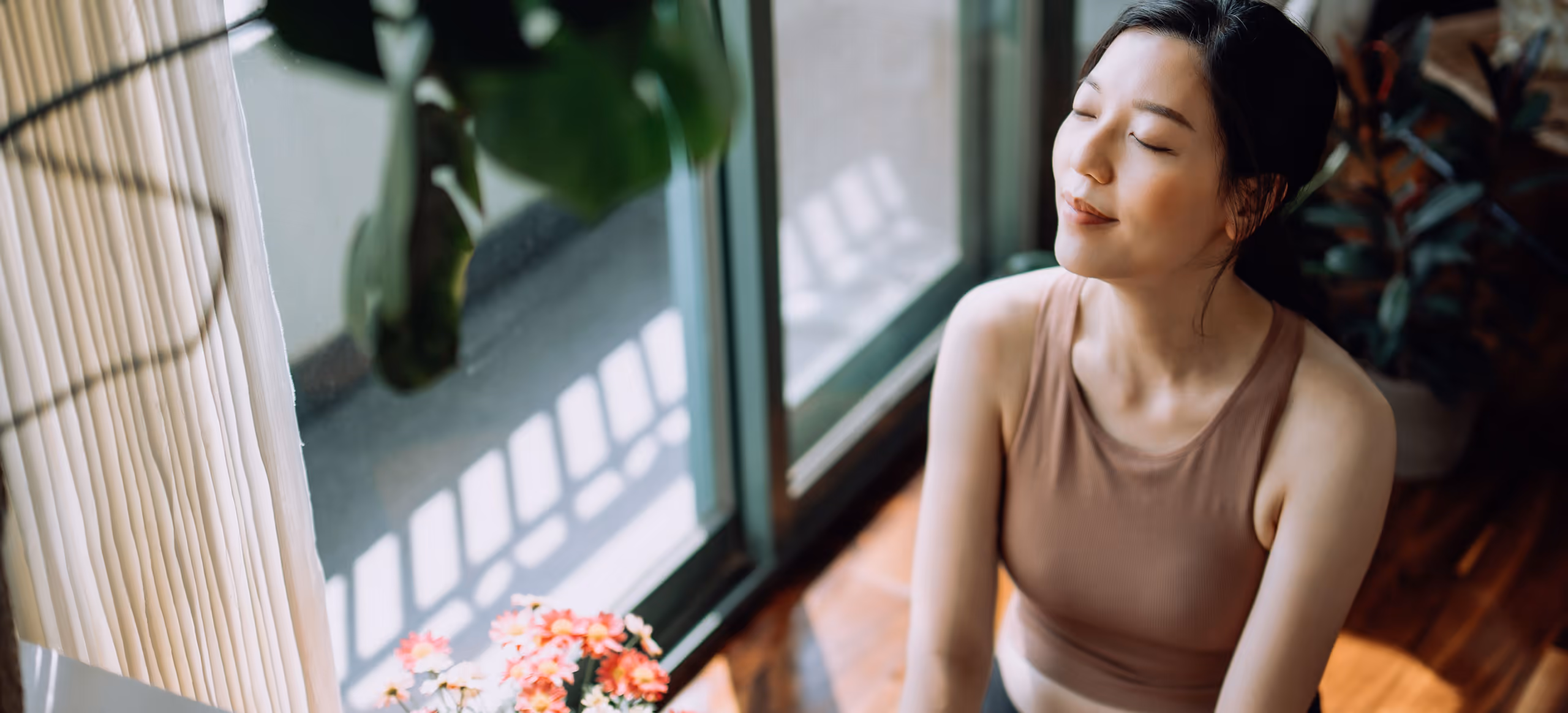 A woman sitting cross-legged by a sunlit window with her eyes closed, plants and flowers around her
