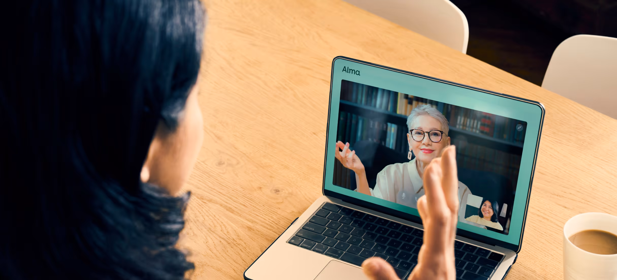 A woman at a laptop on a video call with a therapist, a coffee and phone on the table beside her