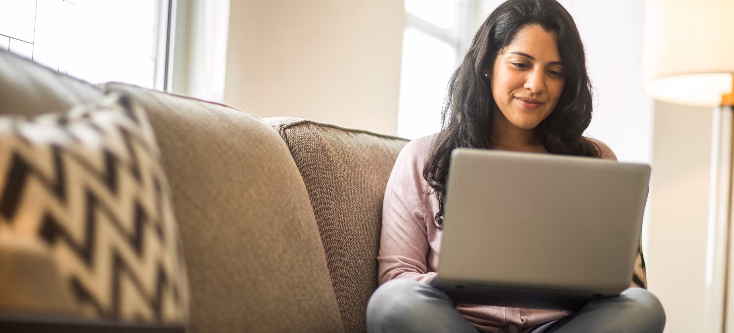 A woman sitting cross-legged on a couch, smiling at a laptop