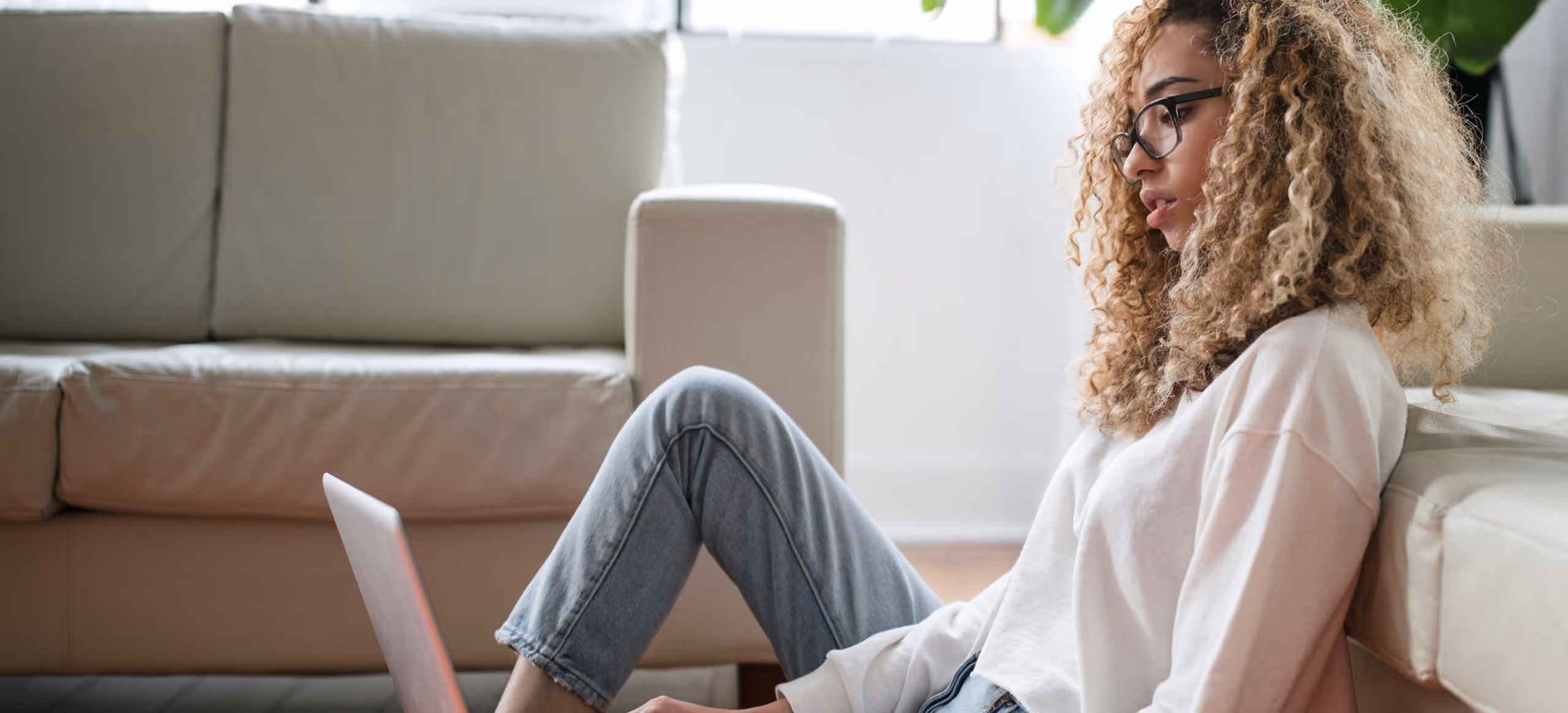 A woman sitting on the floor against a sofa with a laptop on her knees