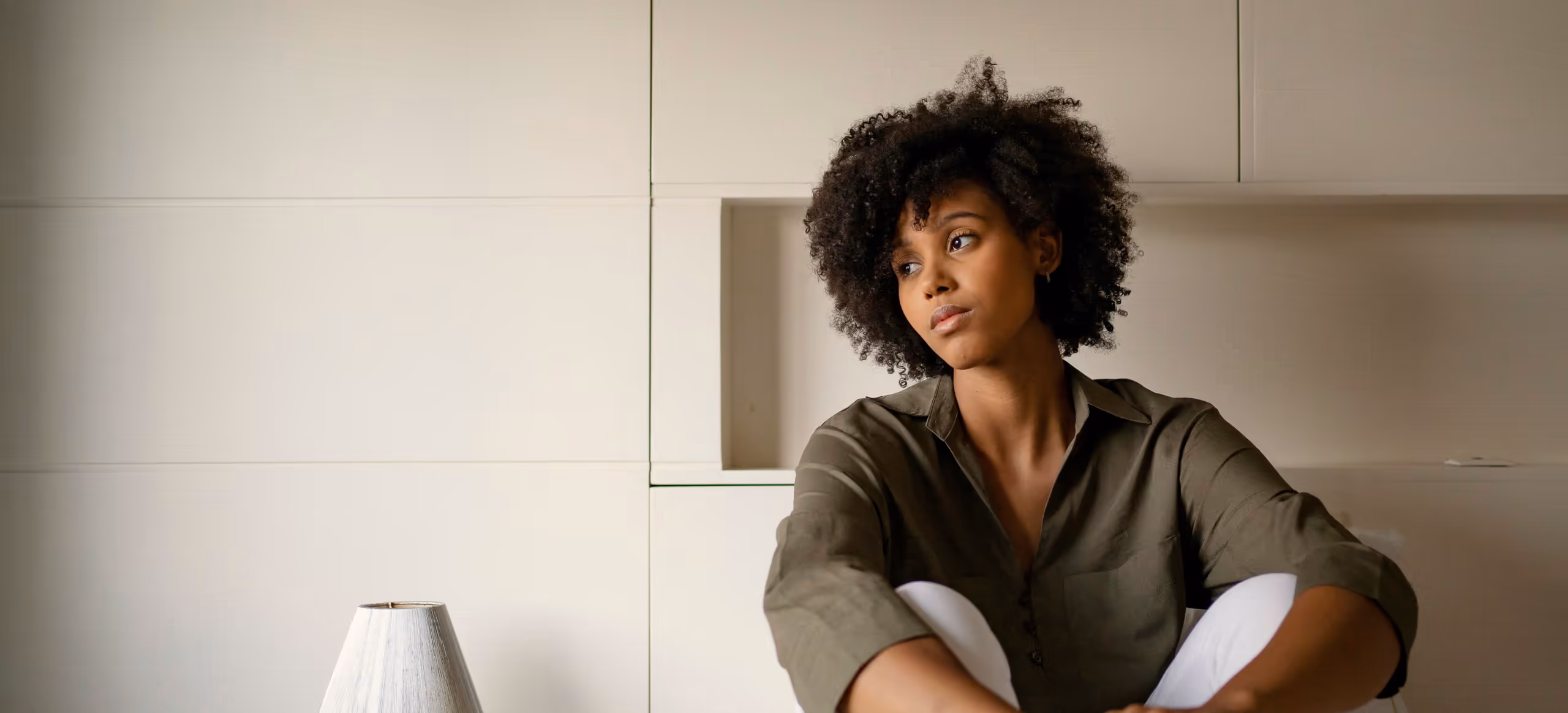 A woman sitting on the edge of a bed with her arms around her knees, looking away in thought