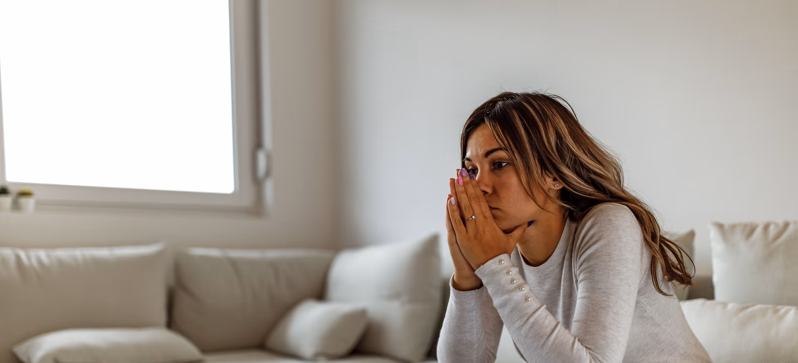 A woman sitting on the edge of a pale sofa with her hands clasped in front of her face